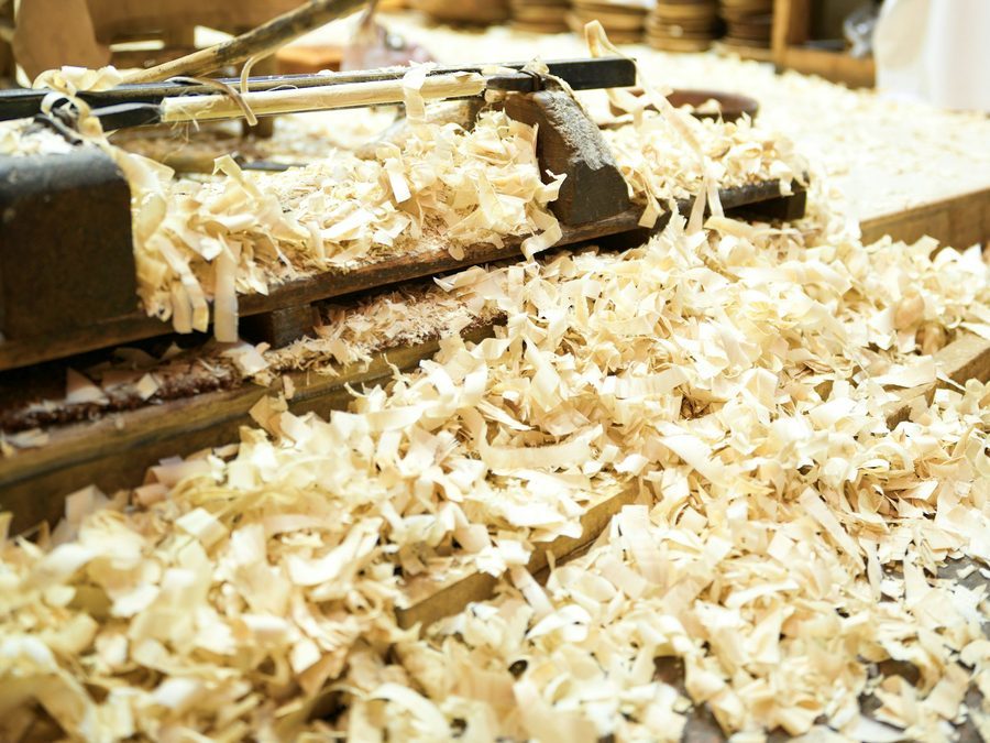 Hobelspäne aus hellem Holz liegen dicht auf einer Werkbank, dahinter ein Handhobel und Regale mit Holzkästen